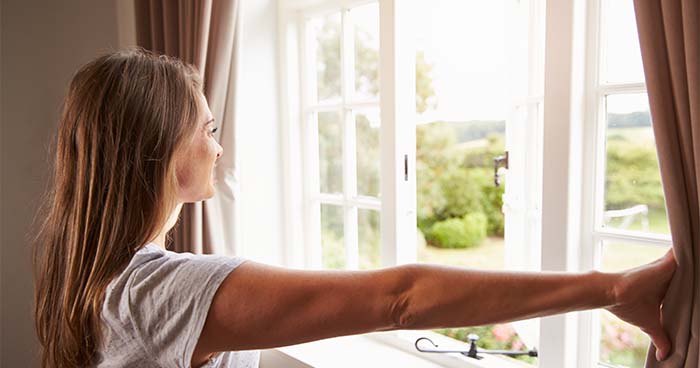 Image: a woman opening curtains to a bright, sunny day. You can save on your electric bill in the winter by opening the curtains during the day to let in natural light and heat.